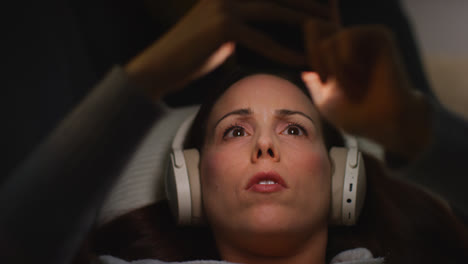 Close-Up-Of-Woman-Wearing-Wireless-Headphones-Lying-On-Sofa-At-Home-At-Night-Streaming-Or-Looking-At-Online-Content-On-Mobile-Phone-1